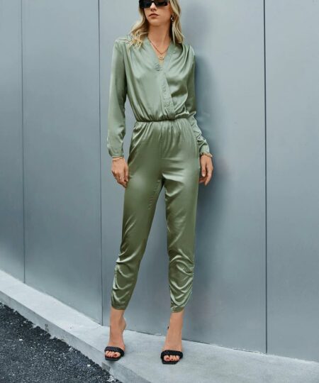 On voit une belle femme blonde qui pose devant un mur gris-bleu et qui porte une combinaison kaki en satin.