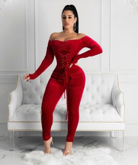 Femme brune avec cheveux attachés et grandes créoles portant une combinaison pantalon moulante en velours rouge avec épaules dénudées et corsage lacé sur le buste avec poches sur les hanches. Elle porte des sandales transparentes à talon sur un tapis en fourrure blanc. Les murs de la pièce sont blancs avec des moulures et Il y a un canapé en simili cuir à capiton blanc derrière la femme.