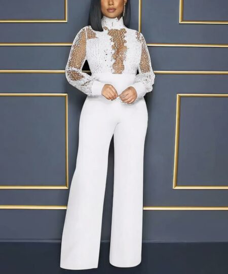 Devant un mur bleu avec moulures dorées, une femme portant une combinaison blanche très élégante avec pantalon droit avec dentelle sur le bustier et le col avec des manches longues transparentes avec sequins à reflets.