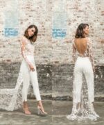 Femme posant devant un mur en pierre avec une combinaison de mariage blanche et en dentelle transparente.
