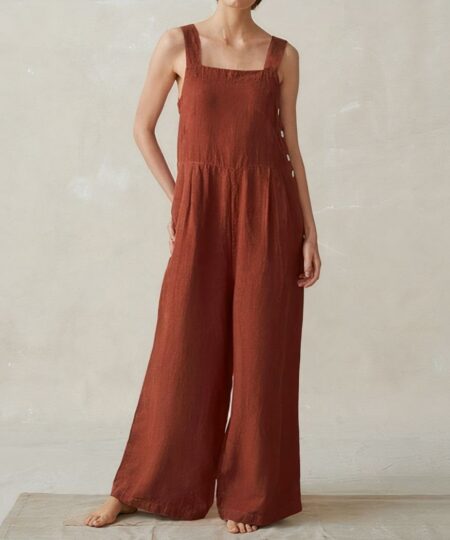 On voit le corps d'une femme sur fond clair qui porte une combinaison couleur terracotta. La combinaison a les jambes larges et des bretelles larges.