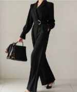 On voit une jeune femme brune très élégante et élancée qui porte une combinaison tailleur smoking noire avec de légères rayures blanches. Le vêtement est joliment ceinturé. La femme porte aussi un petit sac à main noir.