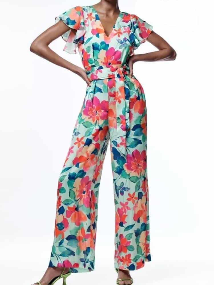 Sur fond blanc, une femme de face avec les mains sur les hanches, portant une combinaison pantalon fond blanc avec fleurs multicolores aux jambes larges , col en v et manches à volants.
