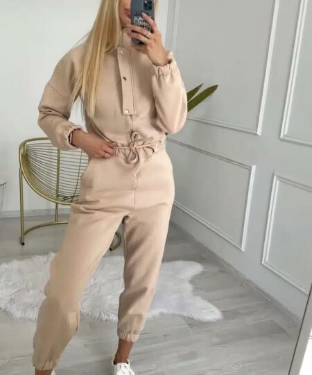 On voit une jeune femme blonde qui porte une combinaison beige en matière polaire. Les manches sont longues. Elles est dans une pièce cossue : les murs sont blancs avec des moulures. Il y a un fauteuil et un tapis en peau de mouton, sur du parquet gris.