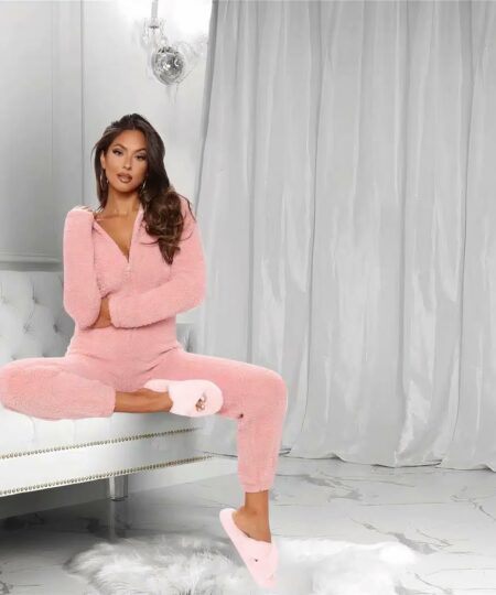 On voit une femme brune aux cheveux longs. Elle porte une combinaison en polaire rose et des chaussons en pilou-pilou. Elle est assise sur un canapé gris près d'un rideau.