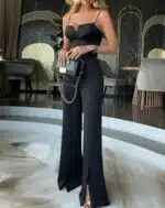 On voit une jeune femme élancée qui porte une combinaison noire à fines bretelles. Le bas du pantalon est fendu. Elle porte à la main un petit sac à maon, et elle se trouve dans un endroit qui ressemble à un hall d'hôtel prestigieux.