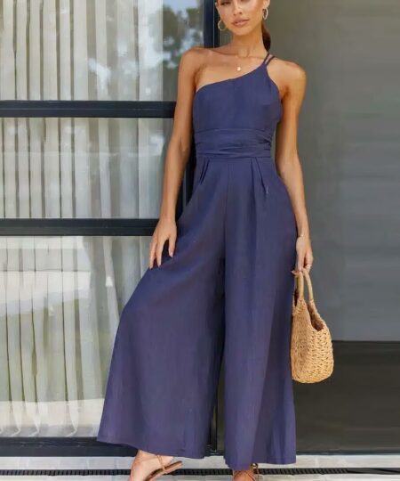 On voit une femme brune élancée sur le pas d'une baie vitrée. Elle porte une combinaison bleu marine en lin avec le haut asymétrique. Elle porte aussi un petit panier en osier estival.