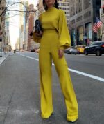On voit une femme brune élancée qui se trouve au milieu de la rue, dans une grande ville qui ressemble à New-York. La jeune femme porte une combinaison jaune manches longues et un sac qu'elle porte à la main.