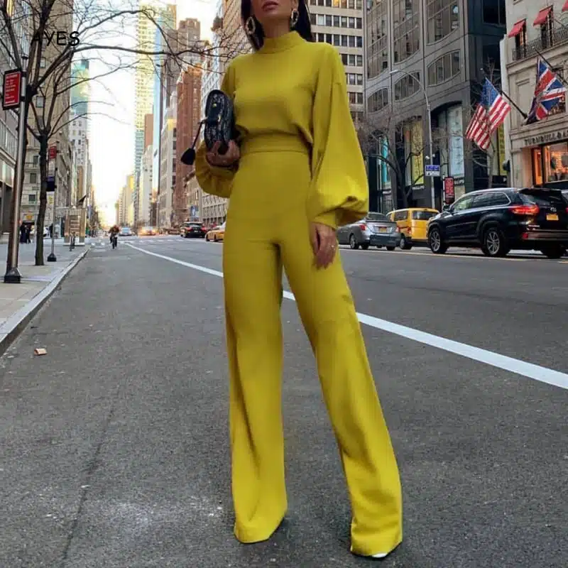 On voit une femme brune élancée qui se trouve au milieu de la rue, dans une grande ville qui ressemble à New-York. La jeune femme porte une combinaison jaune manches longues et un sac qu'elle porte à la main.