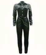 Sur fond blanc, une combinaison pantalon noire à manches longues en simili cuir avec fermeture centrale par boutons pressions argentés et une ceinture à nouer à la taille.