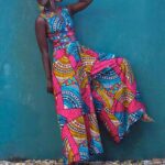 Femme noire devant un mur bleu la jambe levée portant une combinaison pantalon à jambes larges sans manches avec motifs et imprimés africains colorés en rose, bleu et jaune.