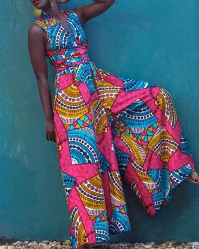 Femme noire devant un mur bleu la jambe levée portant une combinaison pantalon à jambes larges sans manches avec motifs et imprimés africains colorés en rose, bleu et jaune.