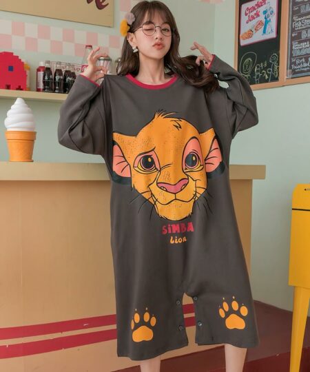 On voit une jeune femme d'origine asiatique qui porte une combinasion courte de pyjama en coton. La combinaison est grise avec la tête de Simba dessus et deux traces de pattes au niveau des genoux. La femme se trouve dans une pièce richement décorée, très pop.