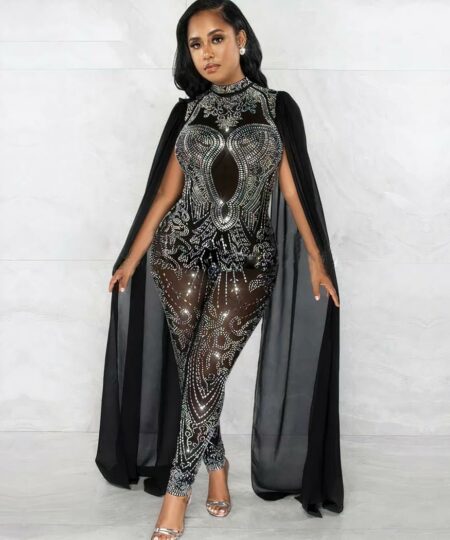 Une femme se tient debout devant un fond dans une combinaison noire à strass avec des manches cape qui descendent jusqu'au sol.