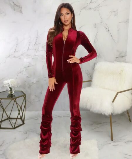 Femme aux cheveux longs châtains portant une combinaison pantalon vintage en velours rouge avec manches longues et fermeture zippée centrale. Il y a un effet drapé sur le bas des jambes au niveau des mollets. Le décor est marbré de gris avec en arrière plan: une table basse filaire noire en métal avec un vase rond en verre transparent avec un petit bouquet de fleur blanche et un fauteuil bas en fourrure blanche.