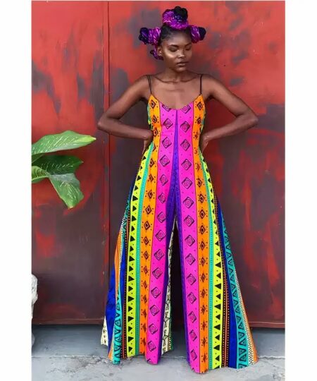 Une femme noire avec chignons et rubans violets dans les cheveux portant une combinaison d'été ample avec un col en V et un pantalon évasé aux motifs tribaux à bandes multicolores. Elle pose devant un mur rouge tacheté a coté d'une plante les mains sur les hanches.