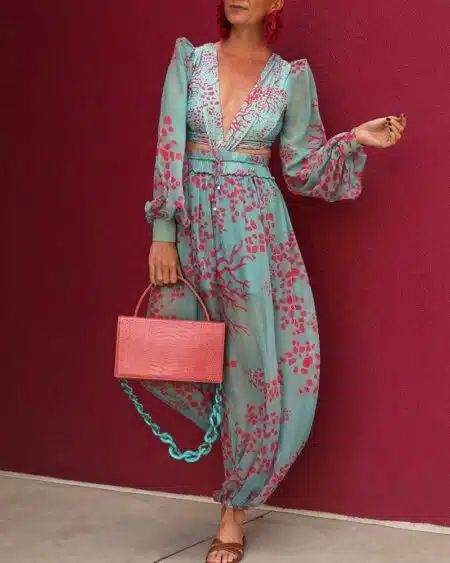 Femme devant un mur rouge portant une combinaison sarouel élégante transparente bleu avec imprimé floral rose, avec manches longues et col en V plongeant. La femme porte un sac en cuir rose et des sandales plates marrons.