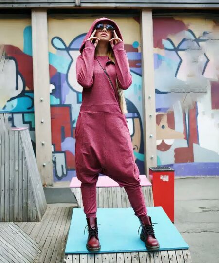 Femme avec lunettes de soleil portant une combinaison sarouel streetwear rose à manches longues avec capuche avec bottines esprit Doc Martens. Elle est dans à l'extérieur avec un mur graffiti derrière elle.