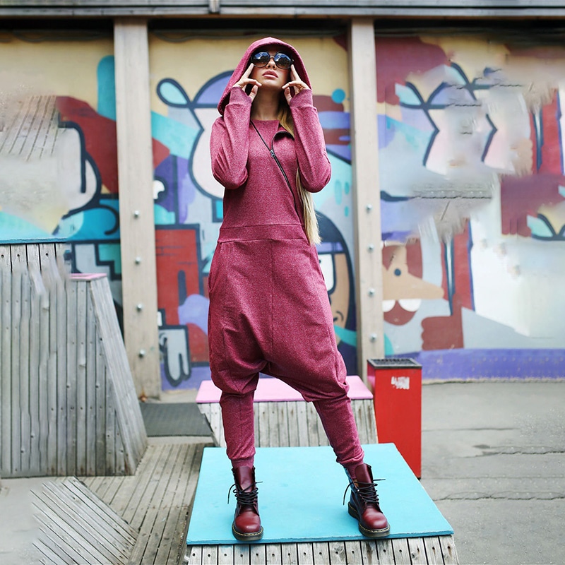Femme avec lunettes de soleil portant une combinaison sarouel streetwear rose à manches longues avec capuche avec bottines esprit Doc Martens. Elle est dans à l'extérieur avec un mur graffiti derrière elle.