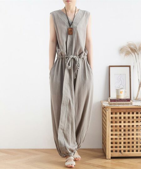 Dans une pièce à la décoration minimaliste composée d'un guéridon en rotin et d'un cadre en bois, on voit une femm qui porte une combinaison en lin grise nouée à la taille. Elle est très élégante.