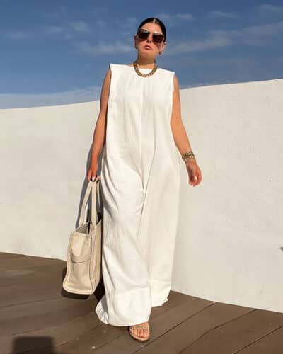 femme portant une combinaison blanche sans manches chic et décontractée marche devant un mur blanc sur un sol en bois en portant un sac des et des lunettes de soleil tout en gardant les bras le long du corps
