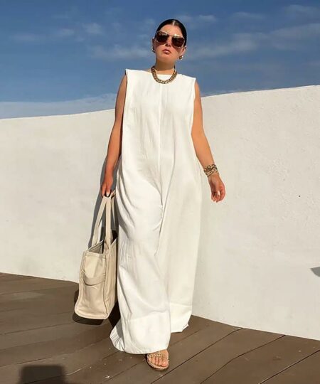 femme portant une combinaison blanche sans manches chic et décontractée marche devant un mur blanc sur un sol en bois en portant un sac des et des lunettes de soleil tout en gardant les bras le long du corps