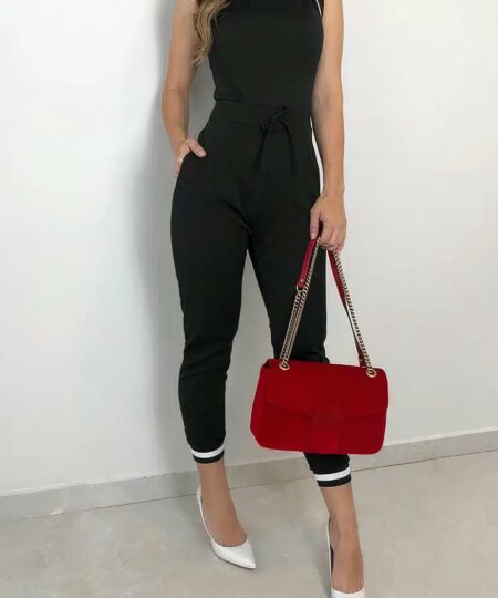 femme portant une combinaison longue noire portant un sac à main rouge et des talons blancs tout en posant devant un mur blanc.