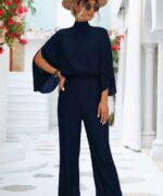 femme blonde posant sur un sol blanc devant des escaliers blanc et des fleurs rouge en portant une combinaison longue chic col montant pour femme bleu marine en portant un chapeau, des lunettes et des talons