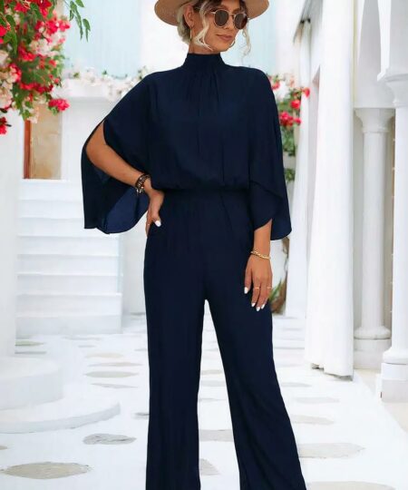 femme blonde posant sur un sol blanc devant des escaliers blanc et des fleurs rouge en portant une combinaison longue chic col montant pour femme bleu marine en portant un chapeau, des lunettes et des talons