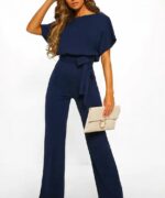 femme portant une combinaison manches courtes chic taille haute bleu marine pose sur fond uni en portant un sac à main blanc et des talons