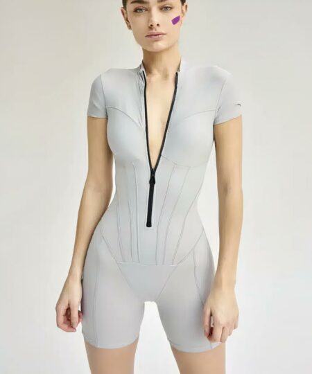 femme portant une combishort décontractée moulante manches courtes grise regarde en face d'elle les bras le long du corps avec la fermeture baissée devant un mure de couleur unie