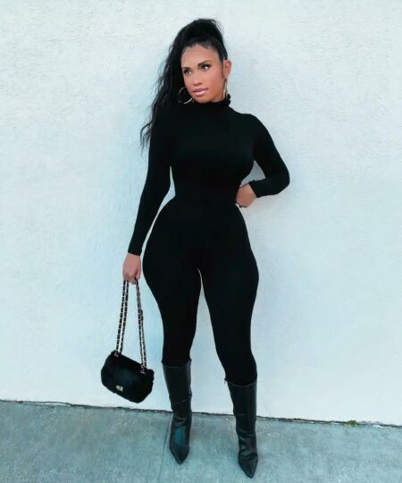 femme brune portant une combinaison moulante manches longues a col roulé noire et un sac à main devant un mur blanc en portant des talons