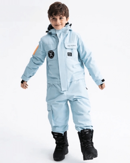 Combinaison de ski enfant épaisse et imperméable avec une capuche portée par un petit garçon avec des bottes noires sur fond gris