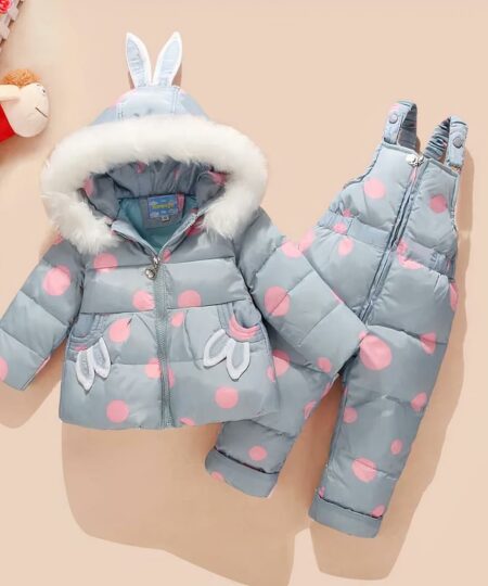 Combinaison de ski enfant avec des oreilles de lapin et motif à pois sur fond beige avec des jouets en haut à gauche