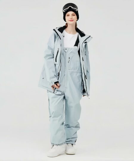 Combinaison de ski femme avec pantalon imperméable et coupe-vent portée par une femme avec un bonnet noir et des bottes blanche sur fond gris