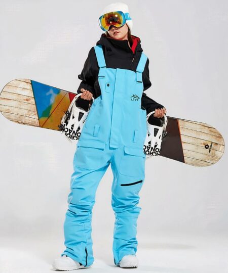 Combinaison de ski femme salopette imperméable et coupe-vent portée par une femme avec un snowboard derrière elle sur fond gris