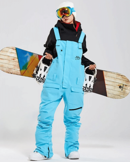 Combinaison de ski femme salopette imperméable et coupe-vent portée par une femme avec un snowboard derrière elle sur fond gris