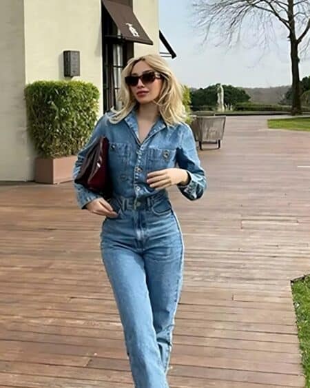 Femme blonde dehors portant une combinaison en jean bleue, des lunettes de soleil et un sac à main