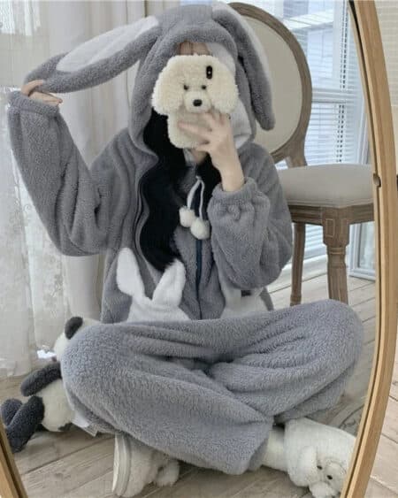 femme en combinaison pyjama en forme de lapin grise et blanche qui est assise sur le sol en tailleur devant un miroir et elle se prend en photo avec son téléphone portable.