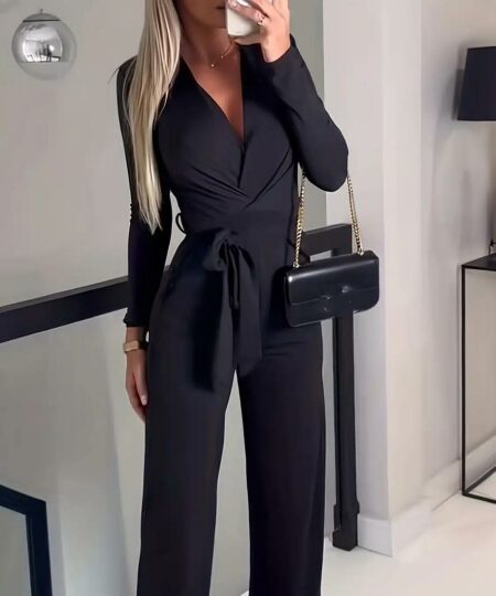 on voit une femme blonde dans un couloir, elle est habillée en combinaison pantalon noire à manches longues et se prend en photo avec son portable elle a un sac à main noir