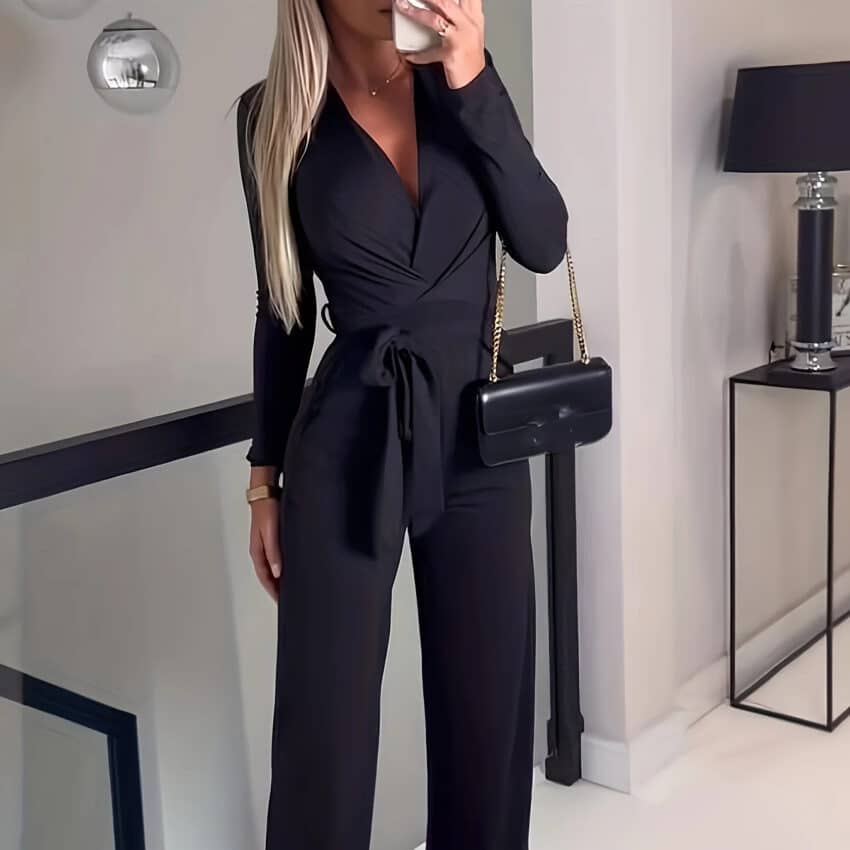 on voit une femme blonde dans un couloir, elle est habillée en combinaison pantalon noire à manches longues et se prend en photo avec son portable elle a un sac à main noir