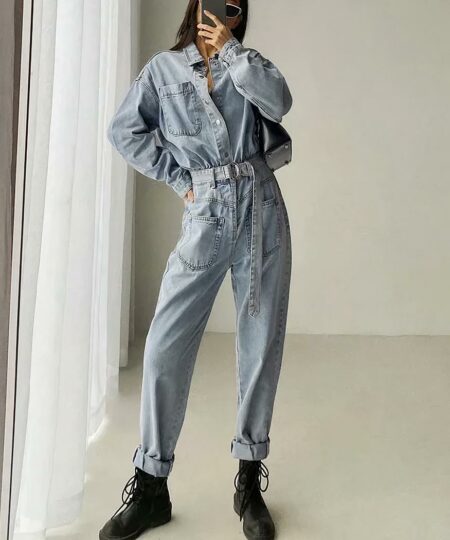 on voit une femme debout dans une pièce claire elle porte une combinaison pantalon en jean clair et des chaussures noires