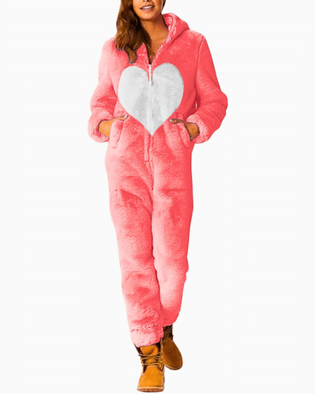 on voit une femme châtain debout les mains dans les poches de sa combinaison pyjama rose avec un grand cœur central blanc elle porte des chaussures marrons